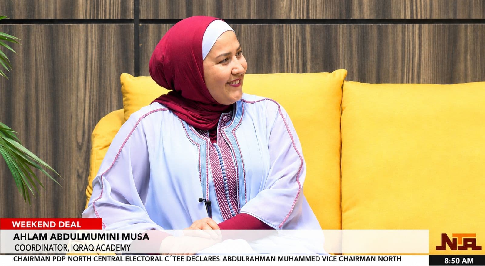 Ahlam Musa, seated on a yellow sofa in front of a wooden wall, smiling and turned slightly away from the camera. It is a still from a television interview, with the station’s caption bar and news ticker along the foot of the picture.