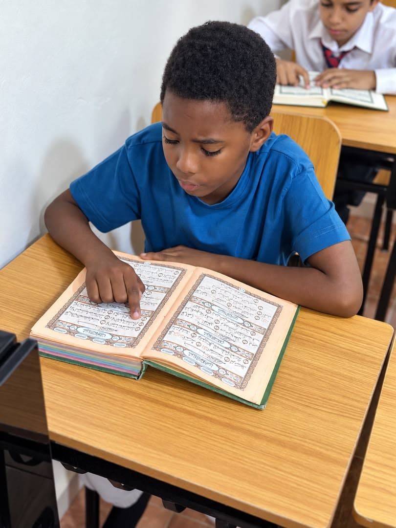A boy in a blue shirt at a wooden desk, reading from a large decorated Qur'an, another student at the desk behind him.