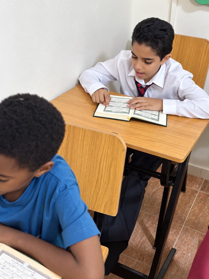 Two students at their desks, one in a white shirt and red tie, reading from a small muṣḥaf.