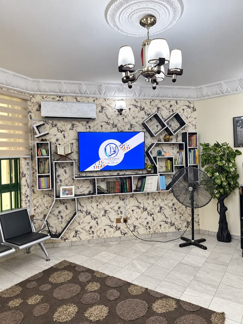 The academy's front room: a screen on a patterned wall showing the Elite Iqraa Academy logo, shelves, a bench and a rug.
