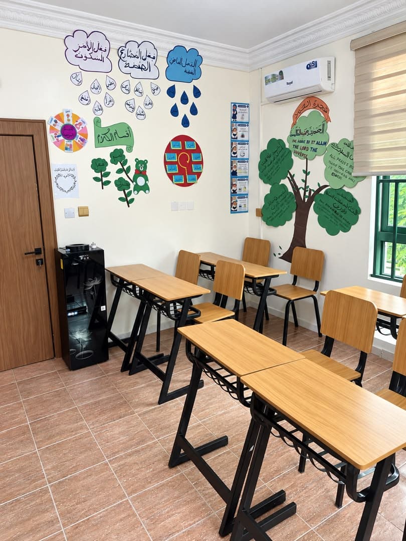 A classroom wall covered with Arabic learning charts, beside wooden desks, a water dispenser and a paper tree.