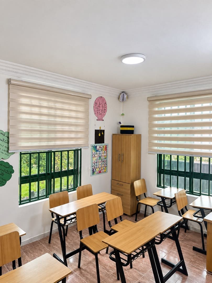 A bright classroom with pale walls, striped blinds, a wooden cupboard and wooden desks.