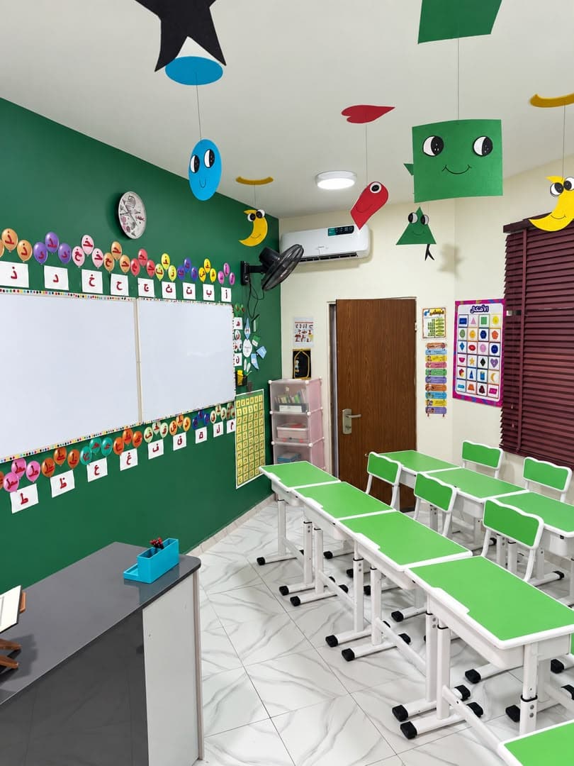 A classroom for young children: a green wall, an Arabic alphabet frieze, a whiteboard, small green desks and paper shapes hanging from the ceiling.