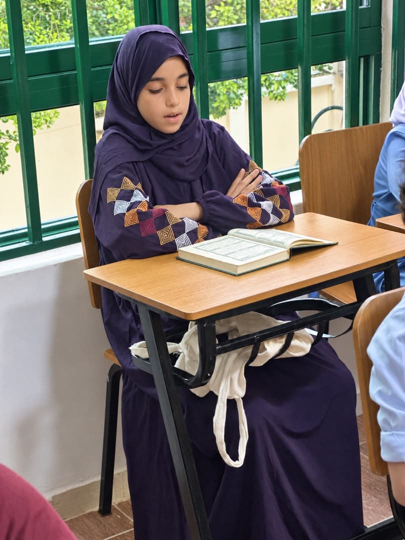 A student in a plain purple hijab and an embroidered gown, reading an open muṣḥaf at a desk beside a tall window.